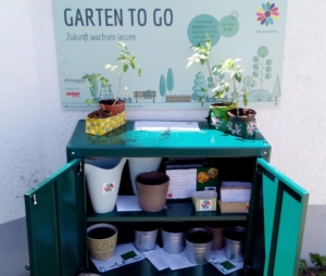Foto des offenen Regals mit Töpfen, Gießkannen und Anleitung, darüber hängt das Schild für den Garten to go