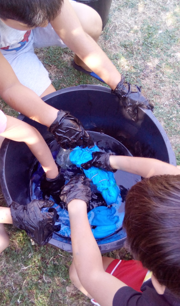 Foto von mehreren Kinderhänden mit Plastik-Handschuhen. Sie tauchen ein T-Shirt in Wasser mit blauer Stoff-Farbe, um es zu färben. (Foto: Katharina Gebel)