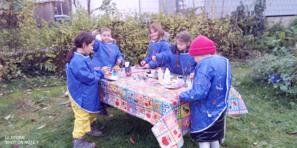 Foto von Kindern mit Malkitteln an einem Tisch im Freien. Sie bemalen gerade Vogelfutter-Kästchen. (Foto: Katharina Gebel)