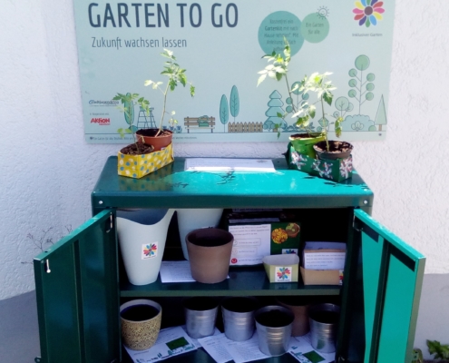 Foto des offenen Regals für den Garten to go mit verschiedenen Pflanztöpfen, Gießkannen und Anleitungen zum Ansäen. Auf dem Regal stehen Tomaten-Pflanzen zum Mitnehmen und darüber hängt das große Schild des Garten to go. (Foto: Katharina Gebel)