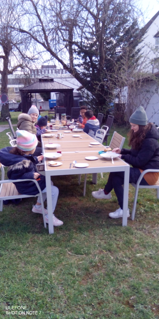 Das Foto zeigt Kinder und Mitarbeiter:innen. Sie sitzen im Garten am Mittags-Tisch. (Foto: Stadtjugendring)