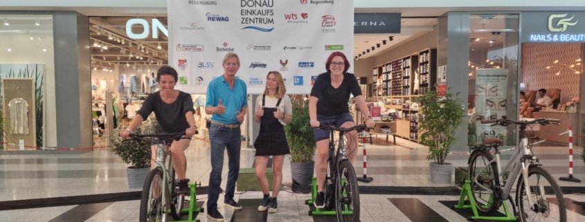 Das Foto zeigt vier Personen im Donaueinkaufszentrum vor dem großen Plakat von "Energie wird Natur". In der Mitte stehen zwei, links und rechts sind zwei Personen auf den Fahrrädern. (Foto: Stefanie Schmid)