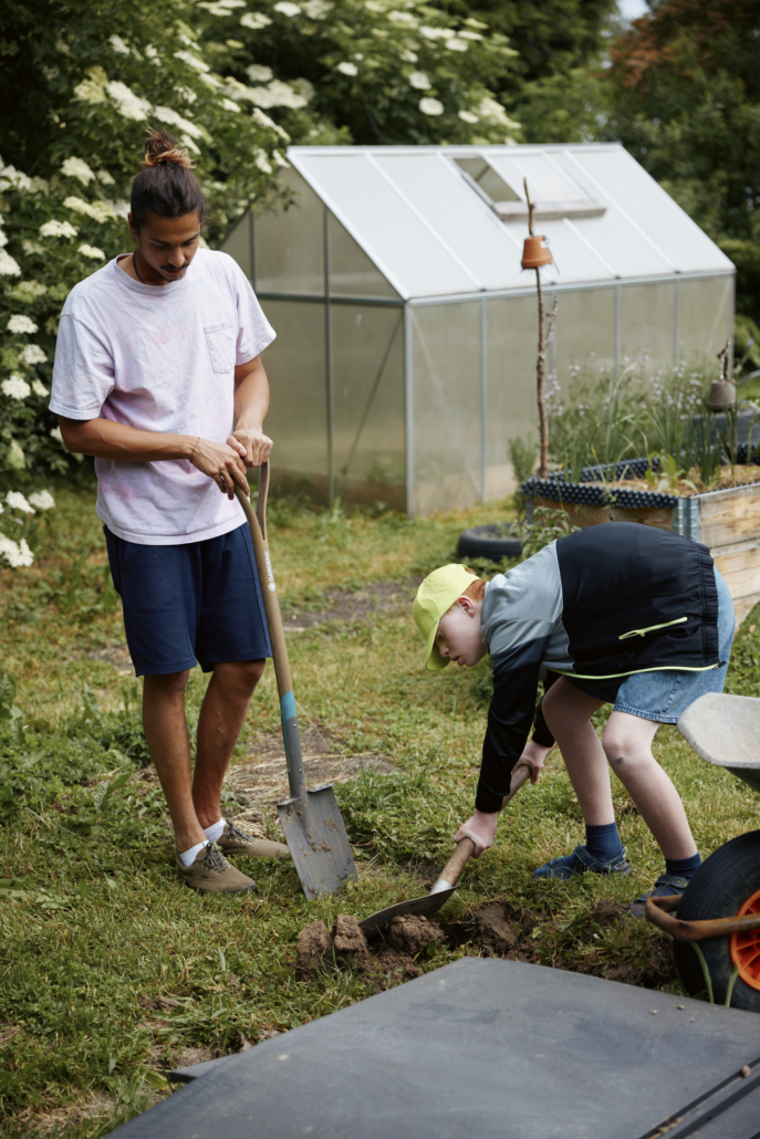 Das Foto zeigt ein anderes Kind und einen Mitarbeiter mit Schaufeln bei der Gartenarbeit. (Foto: ©BJR/Johnke)