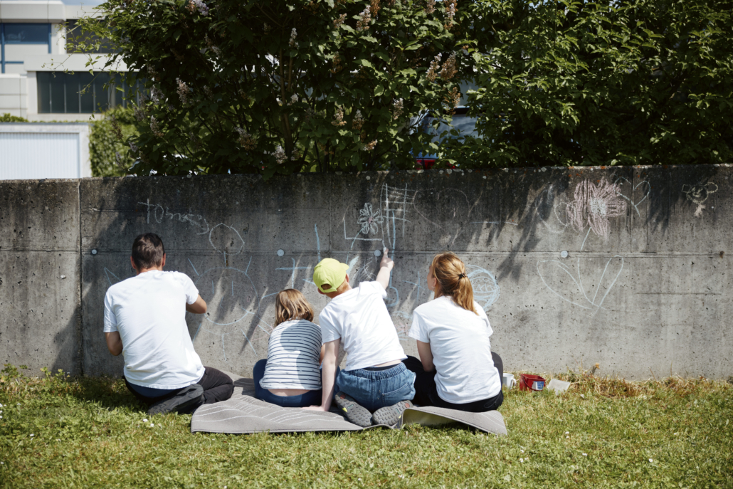 Das Foto zeigt 2 Kinder und 2 Mitarbeiter:innen beim Bemalen unserer Gartenmauer mit Kreide. (Foto: ©BJR/Johnke)
